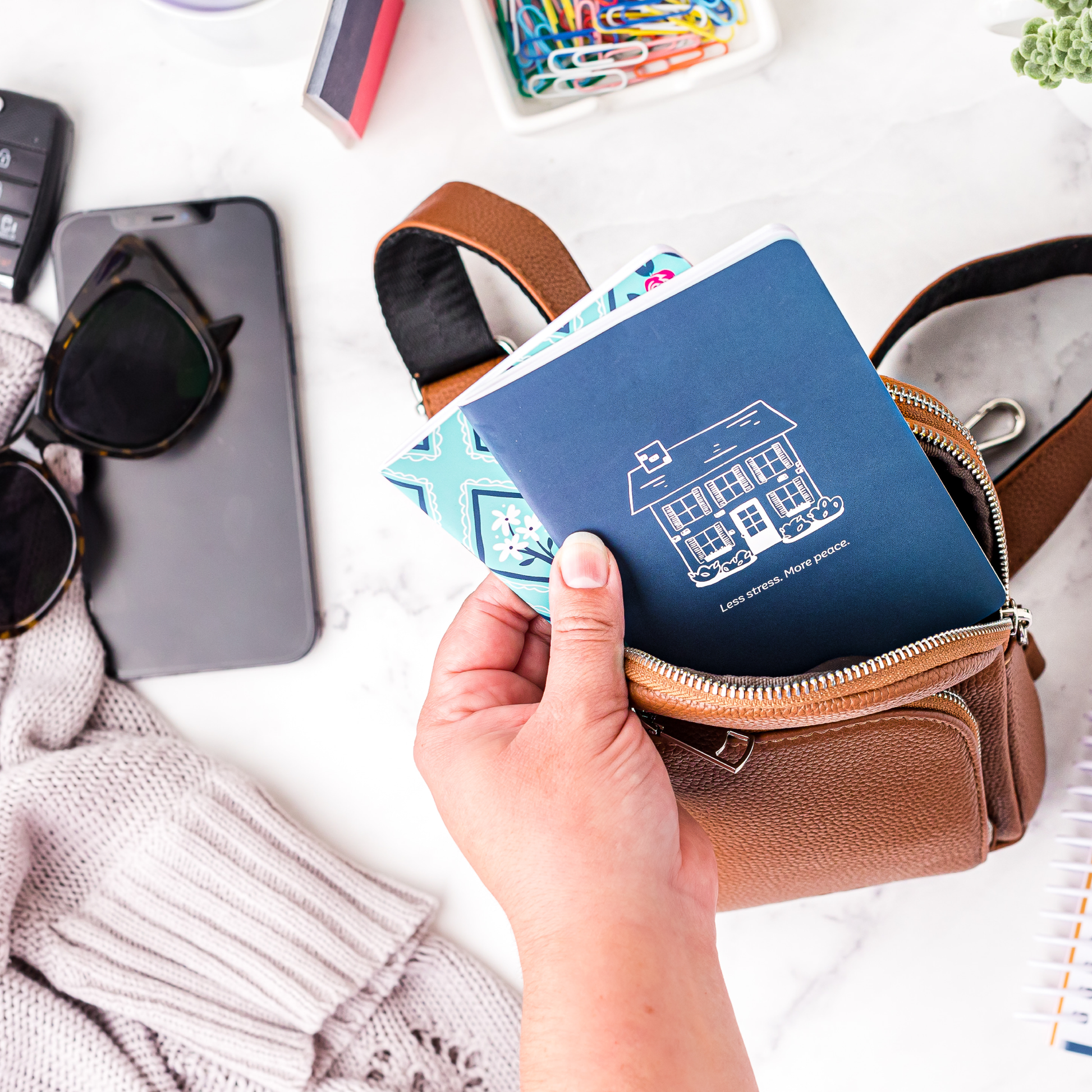 Person putting their Less Stress More Peace mini notebook into a purse, surrounded by travel items