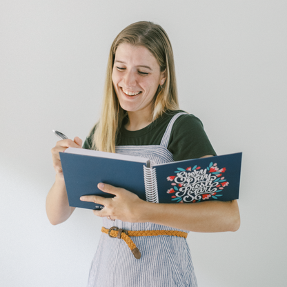 Woman holding a blue planner with decorative cover and pen, smiling against a plain background